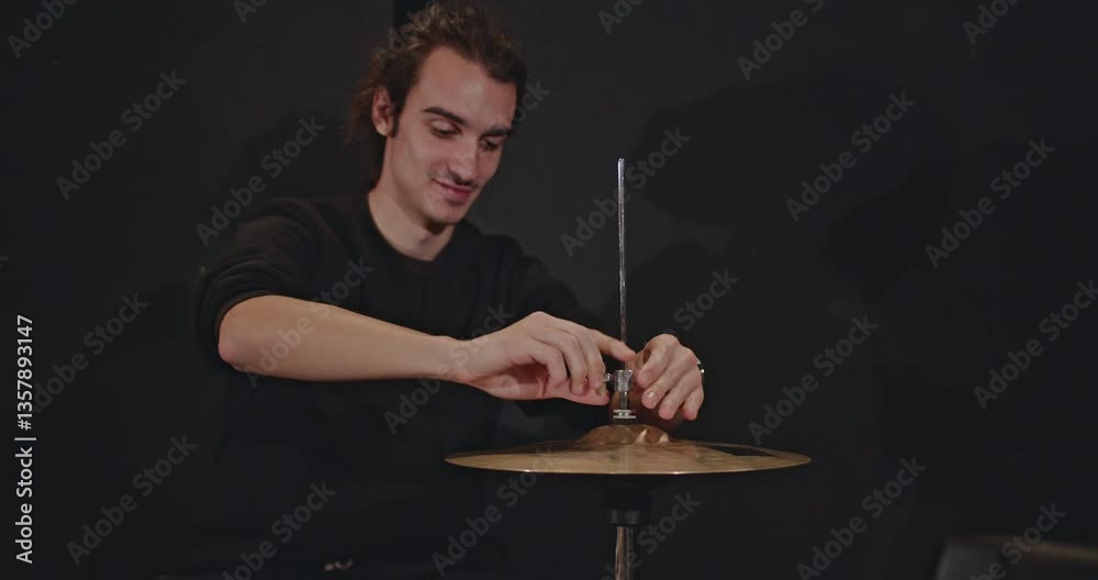 A drummer meticulously adjusts his hi-hat cymbals in a dark studio, preparing for a jazz trio concert. The band practices their music, capturing the essence of live performance.