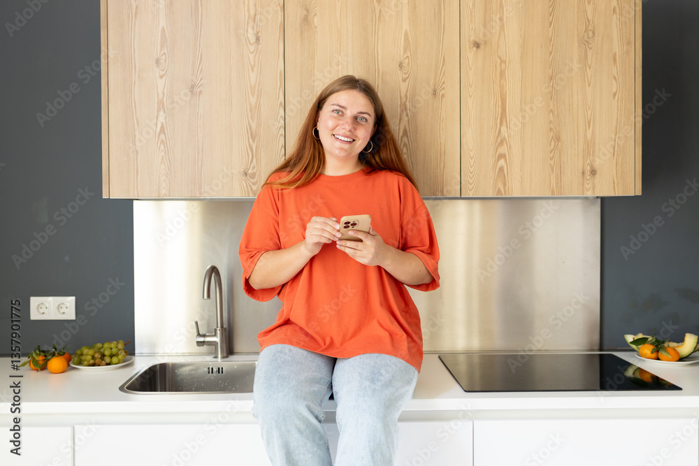 Portrait of smiling housewife is sitting on desk and using phone in modern kitchen. Home, people concept. Copy space. 30s Woman watching recipe on social media, video tutorial how to prepare meal.