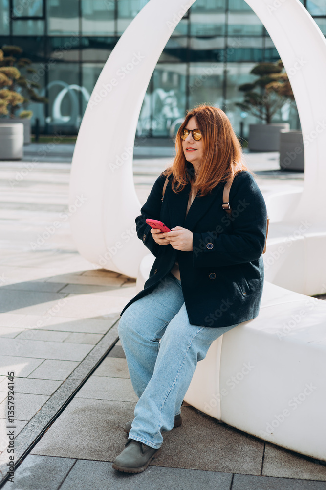 Happy woman chatting on phone and relaxing outdoors. Urban, people concept. Smiling 30s redhead woman text messaging on smart phone at sunny day on the city street, spring time