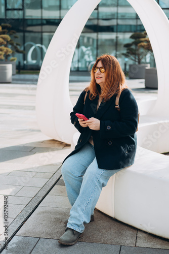 Obraz na płótnie Happy woman chatting on phone and relaxing outdoors. Urban, people concept. Smiling 30s redhead woman text messaging on smart phone at sunny day on the city street, spring time w salonie