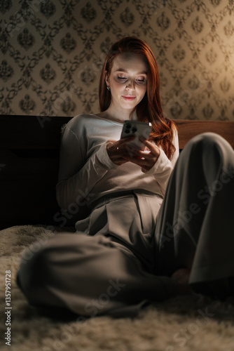 Vertical portrait of focused tired woman using smartphone sitting on bed in dark bedroom at night, light from screen illuminating face. Concept of social media addiction, phone overuse, insomnia. photo