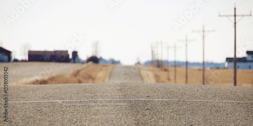 long, straight, rural road stretching into the distance, flanked by open fields, utility poles, and a few scattered buildings