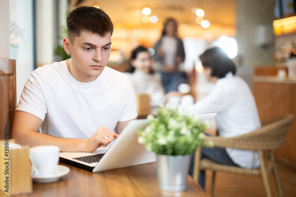 Fototapeta premium Young man working on laptop drinking coffee in cafe