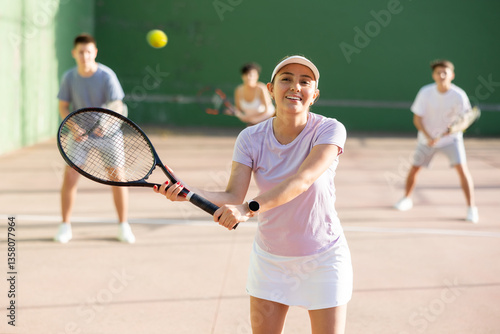 Sporty young Argentinian woman playing popular team match of frontenis at open-air fronton on summer day, ready to hit rubber ball with racquet