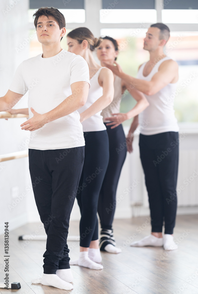 Focused young guy, amateur ballet dancer, practicing basic classical positions at barre during group class under guidance of experienced male choreographer in sunlit studio Focused young guy, amateur ballet dancer, practicing basic classical positions at barre during group class under guidance of experienced male choreographer in sunlit studio