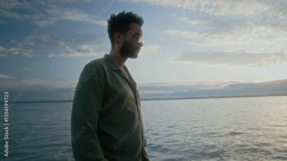 Medium rolling tracking shot of pensive young black man with bare feet walking on sandy lake beach, carrying bright orange daisy flower and smelling it, feeling lonely Medium rolling tracking shot of pensive young black man with bare feet walking on sandy lake beach, carrying bright orange daisy flower and smelling it, feeling lonely