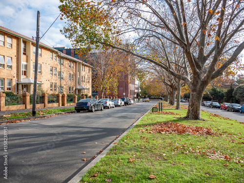 A typical North Melbourne streetscape in Australia during autumn, featuring a wide median nature strip with mature deciduous trees, road with brick apartment buildings and parked cars. photo