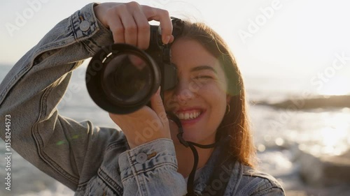 A smiling girl taking a photo with her camera by the sea at sunset. Capturing the beauty of nature with passion and joy