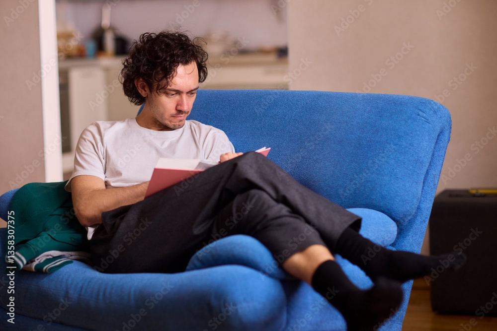 A Young Man Immersed in Reading a Book While Relaxing in a Cozy Living Room Atmosphere A Young Man Immersed in Reading a Book While Relaxing in a Cozy Living Room Atmosphere