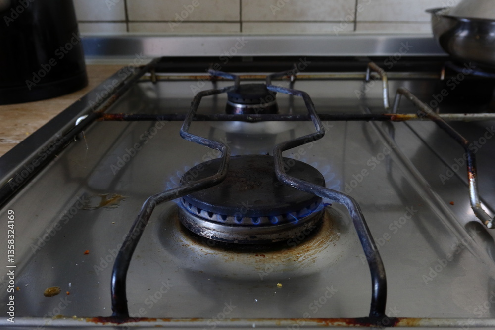 Close-up of a gas stove burner with a visible blue flame. The surface is dirty with food stains and rust, showing a real-life kitchen environment and signs of daily use Close-up of a gas stove burner with a visible blue flame. The surface is dirty with food stains and rust, showing a real-life kitchen environment and signs of daily use