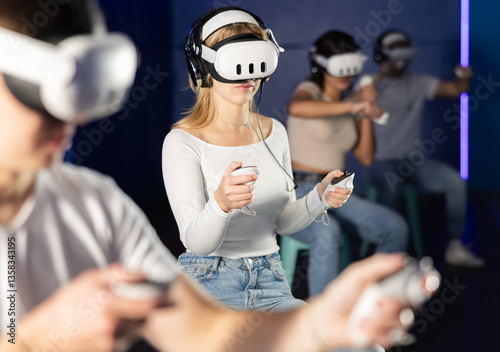 Girl in vr glasses with earbuds and two joysticks in hands is playing an active video game with friends. Female gamer in a virtual reality helmet plunged with friends into simulation