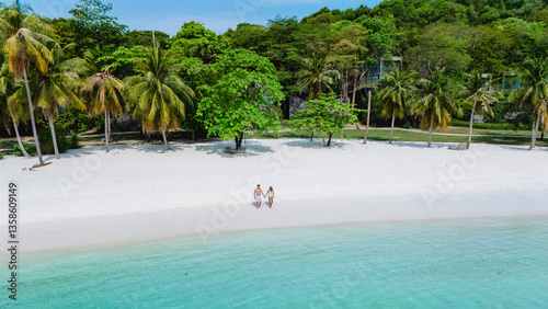 Two people stroll along the pristine white sands of Koh Kham Thailand, framed by swaying palm trees and clear turquoise waters. The vibrant greenery enhances the tropical paradise atmosphere.