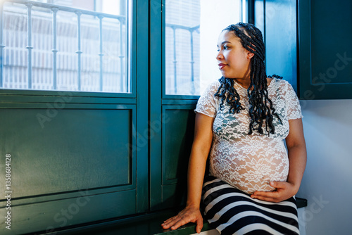Pregnant latin woman sitting next to window holding her belly while thinking about her future
