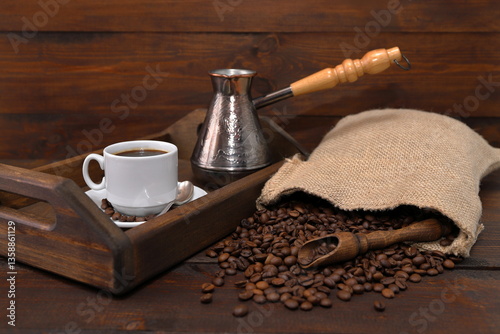 On a brown wooden background there is a wooden tray with a white cup of coffee and a copper turk, next to a scattering of coffee beans from a burlap sack and a wooden scoop with coffee beans. 
