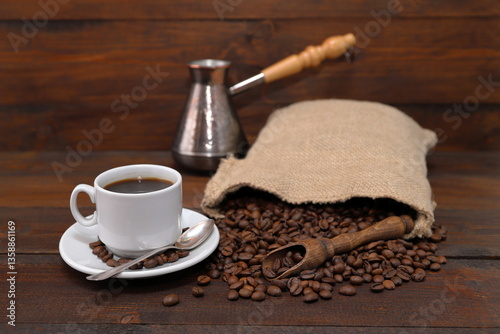 On a brown wooden background there is a white cup of coffee, next to a copper turk, a scattering of coffee beans from a burlap sack and a wooden scoop with coffee beans.