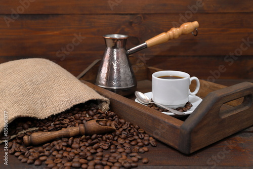 On a brown wooden background there is a wooden tray with a white cup of coffee and a copper turk, next to a scattering of coffee beans from a burlap sack and a wooden scoop with coffee beans. 