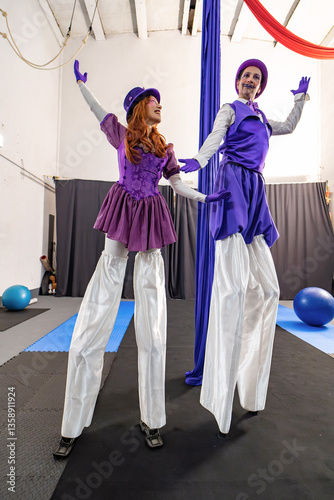 Two stilt walkers wearing purple costumes are performing a show in a circus training room with aerial silks and gym balls