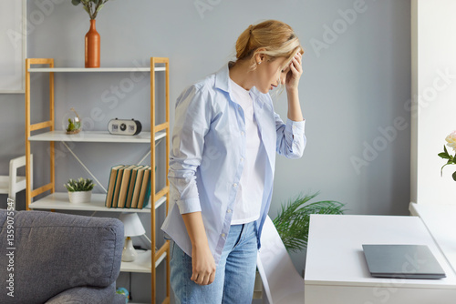 Overworked adult woman suffering from severe headache or migraine, touching forehead, feeling tired after working long hours at computer, standing near desk in living room, having health problems photo
