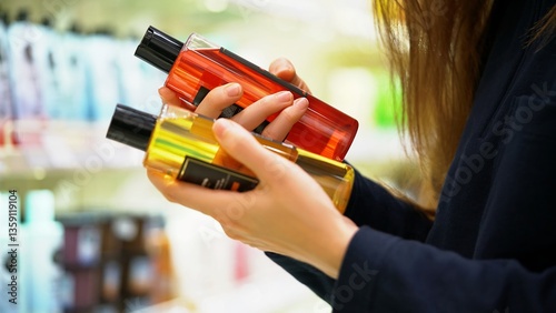 A female customer compares two shampoos, reads the labels, examines the ingredients and choosing for a conscious decision. Close-up of hands holding two different bottles in a cosmetic hair care aisle