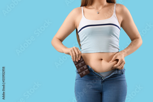 Young woman in tight jeans with chocolate on blue background, closeup. Weight gain concept