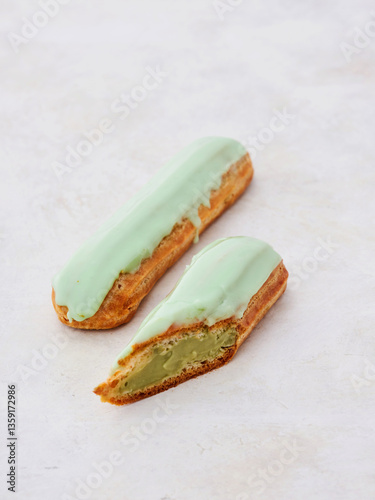 Two delectable eclairs sit on a light surface, one cut in half to reveal a smooth pistachio filling. The bright green glaze adds a vibrant touch, inviting indulgence photo