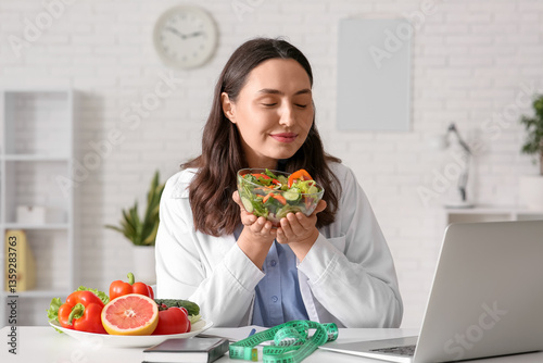 Young female nutritionist with vegetable salad video chatting at table in office
