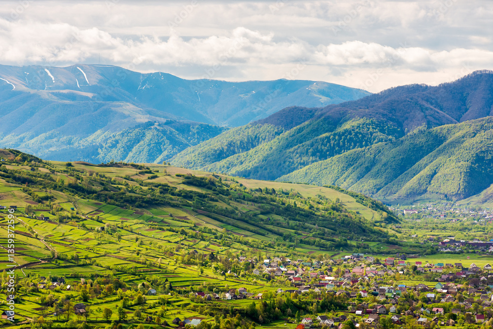 mountainous countryside landscape with kolochava village in the valley. cloudy weather. rural fields and meadows on the forested slopes of a hill. rolling alpine scenery in spring