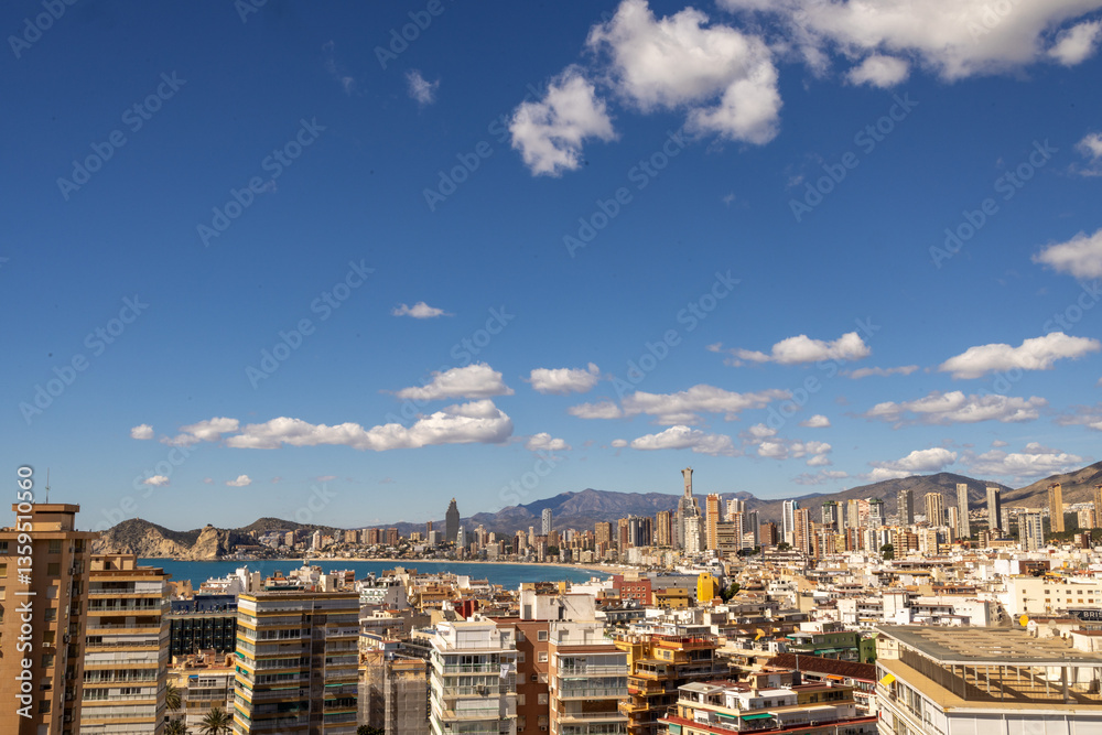 Photo overlooking the old town of Benidorm in Spain showing the West Beach Promenade surrounded by high rise apartments and buildings with the mountains and white clouds in the background