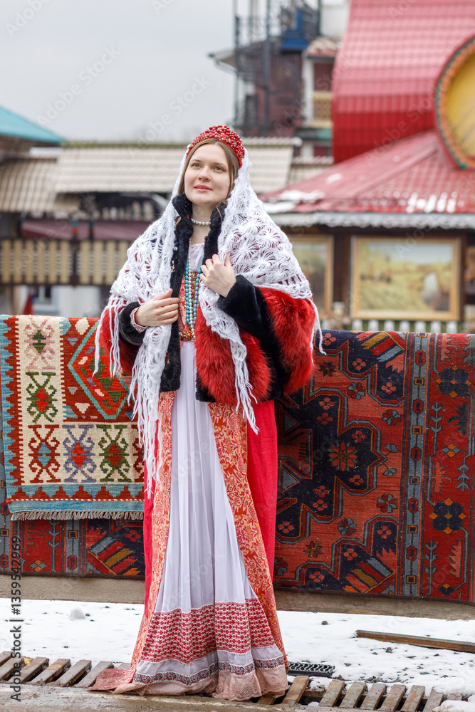 Beautiful Russian girl in red traditional Russian clothes and white shawl near wooden house in spring. Festive farewell to winter