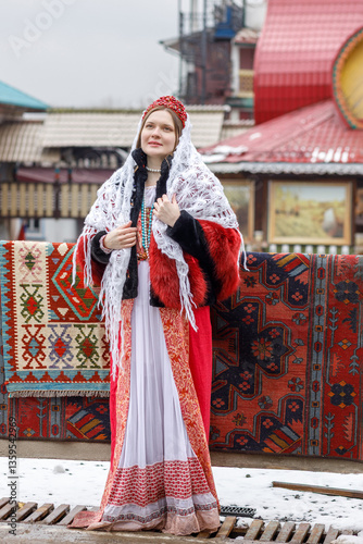 Obraz na płótnie Beautiful Russian girl in red traditional Russian clothes and white shawl near wooden house in spring. Festive farewell to winter w salonie