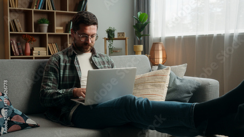 Happy smiling positive Caucasian man male guy at home couch sofa with laptop computer working online internet work typing shopping complete accomplishment done job relax break put hands behind head