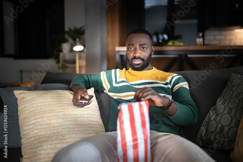 Фотография A serious-looking man sits on a couch watching TV, holding a remote and eating popcorn