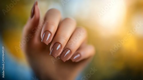 Close-up of a woman's hand showcasing manicured nails with a glossy light brown finish against a soft-focus autumnal background.