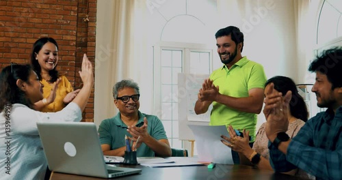 Indian Asian young business people or startup partners celebrating team success with high-fives and cheers while sitting in a meeting with a laptop and documents on the table in a modern cozy office