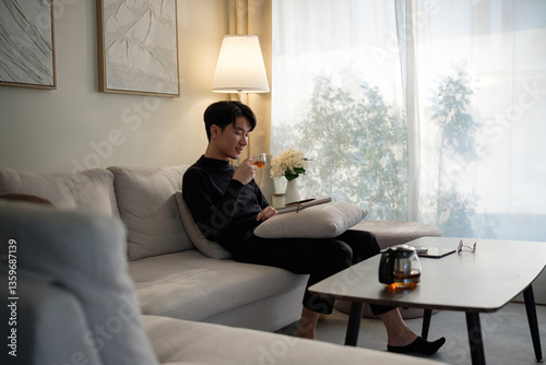 Photos Calm young man in a black sweater sits comfortably on sofa, sipping a warm tea