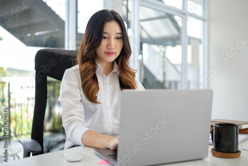 Determined young woman in formal attire focuses on her laptop while seated at a sleek, modern desk. Free Download Determined young woman in formal attire focuses on her laptop while seated at a sleek, modern desk. - GFXLoad