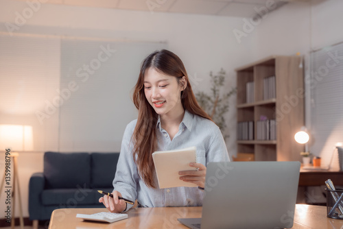 Young asian businesswoman calculating domestic expenses using calculator and taking notes in notebook while working with laptop computer at home office at night