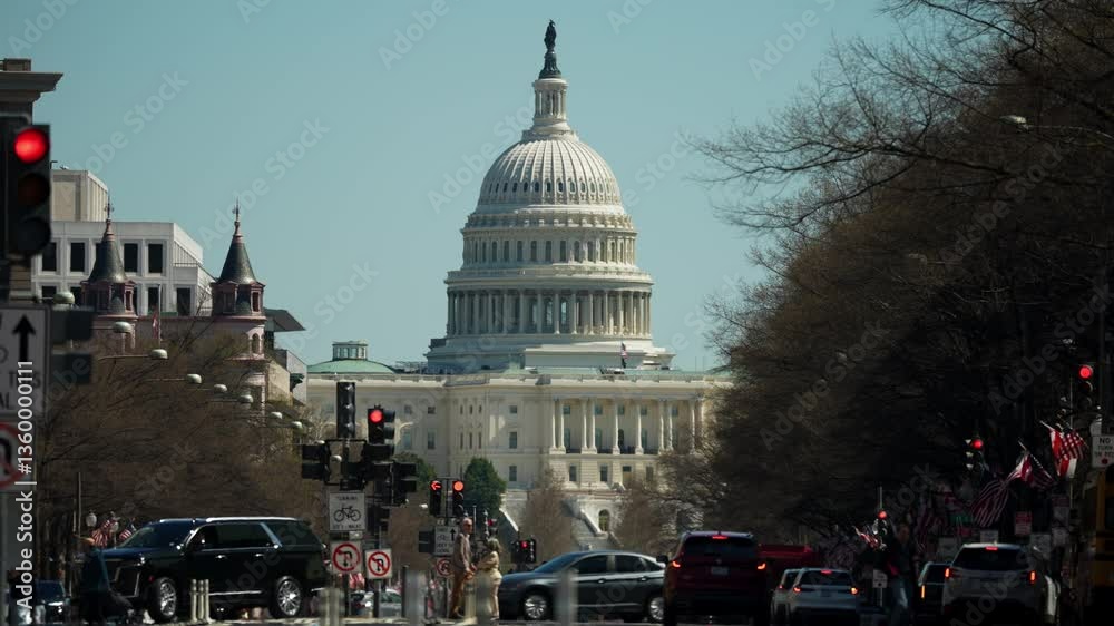 The US Capitol building stands majestically under a clear blue sky, with cars navigating the street and flags adorning the area, creating a vibrant atmosphere.
