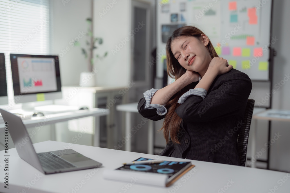 Obraz premium Asian businesswoman massaging her neck while seated at her desk, experiencing discomfort from neck pain after a long day of work in a busy office environment with a laptop