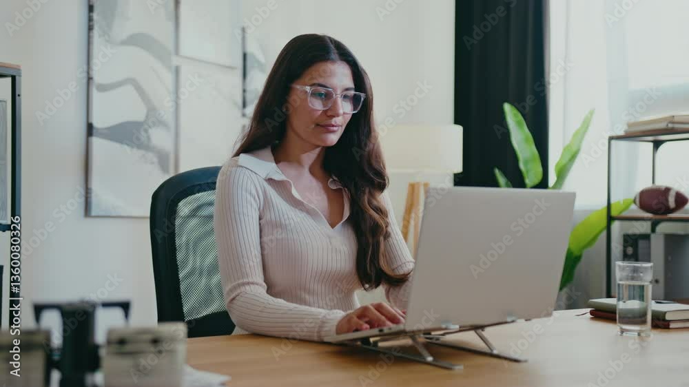 Focused Caucasian woman with long wavy hair and glasses typing on laptop while concentrating on screen. Sitting upright and working in stylish office. Modern workspace with elegant decor. Focused Caucasian woman with long wavy hair and glasses typing on laptop while concentrating on screen. Sitting upright and working in stylish office. Modern workspace with elegant decor.