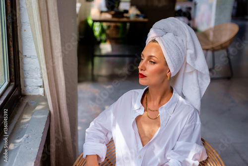 Attractive woman with a towel turban on her head sitting in a chair at the window at natural light