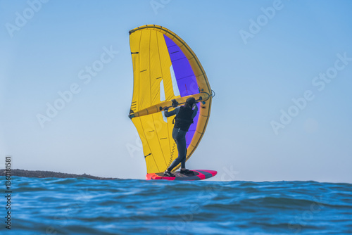 A man wing foils at sea using a brightly coloured yellow and blue hand held inflatable wing, riding a  bright red hydrofoil surfboard. Blue sea and clear blue sky with no clouds.