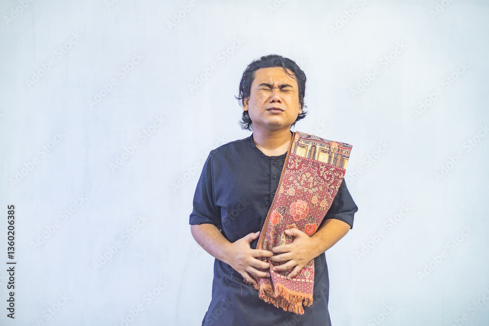 Portrait of young Asian Muslim man holding his stomach because of hunger while fasting. Ramadan and Eid concept.
