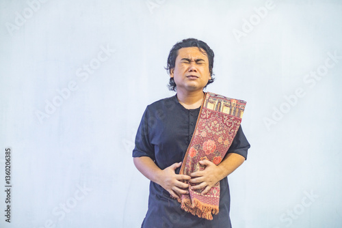 Obraz na płótnie Portrait of young Asian Muslim man holding his stomach because of hunger while fasting. Ramadan and Eid concept. w salonie