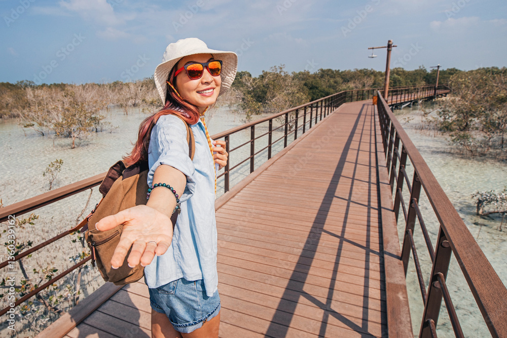 Woman enjoying a sunny day on a wooden boardwalk, extending hand with follow me pose, surrounded by lush mangrove forest coastal park