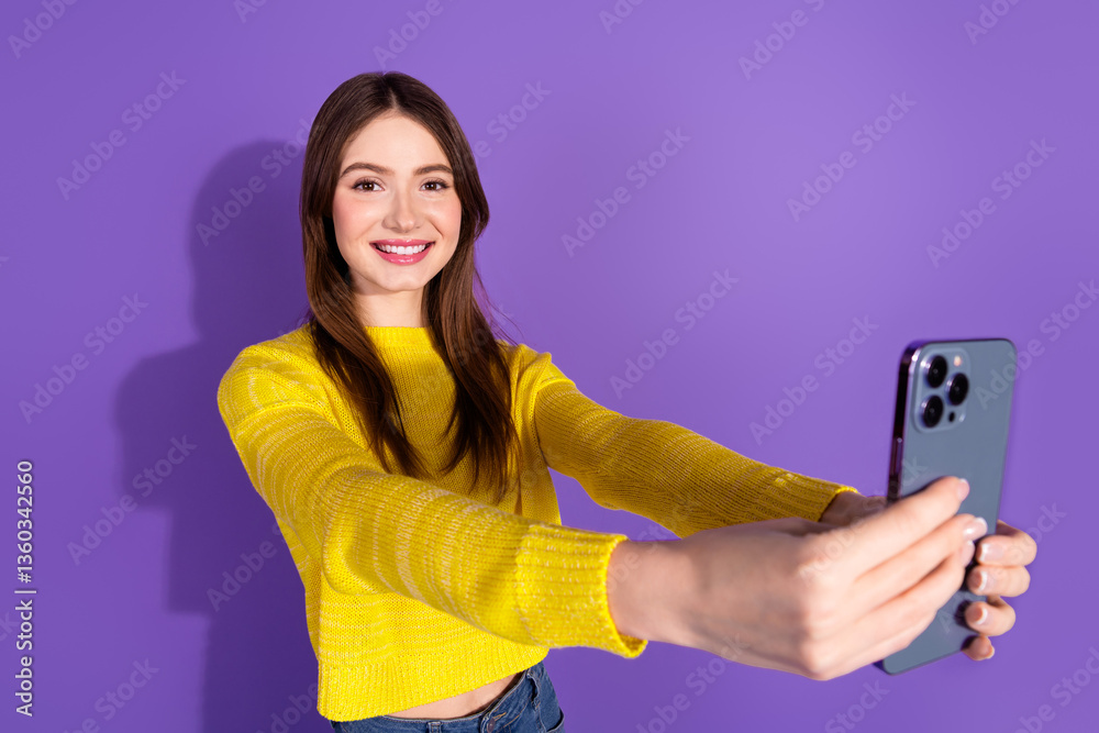 Naklejka premium Smiling young woman in casual yellow sweater taking a selfie against a violet background showcasing cheerful and trendy lifestyle