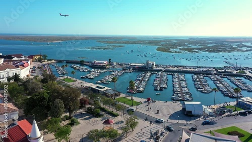 A high-angle view of Faro's marina in Portugal, featuring numerous boats, a scenic promenade, lush greenery, and the beautiful Ria Formosa lagoon with scattered sailboats in the distance.
