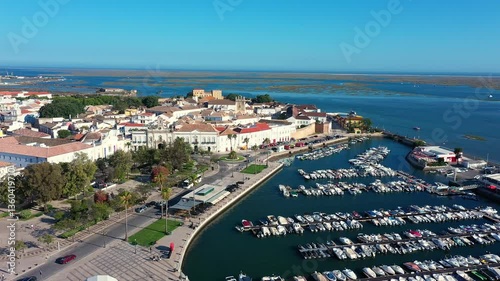 Aerial view of Faro's old town in Portugal, featuring historic buildings, a waterfront promenade, a marina filled with boats, and the stunning blue waters of the Ria Formosa lagoon.