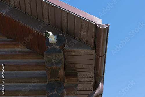 A security camera is mounted on the corner of a wooden house under a brown metal roof. The camera is directed outward, ensuring surveillance and protection. 
