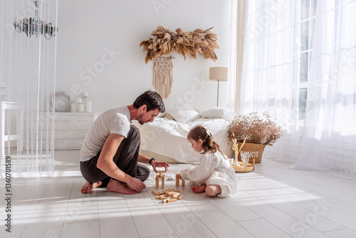 Father and little daughter are sitting on the floor in a cozy bedroom, playing together and building a tower with wooden blocks, enjoying quality time and creating a loving family bond